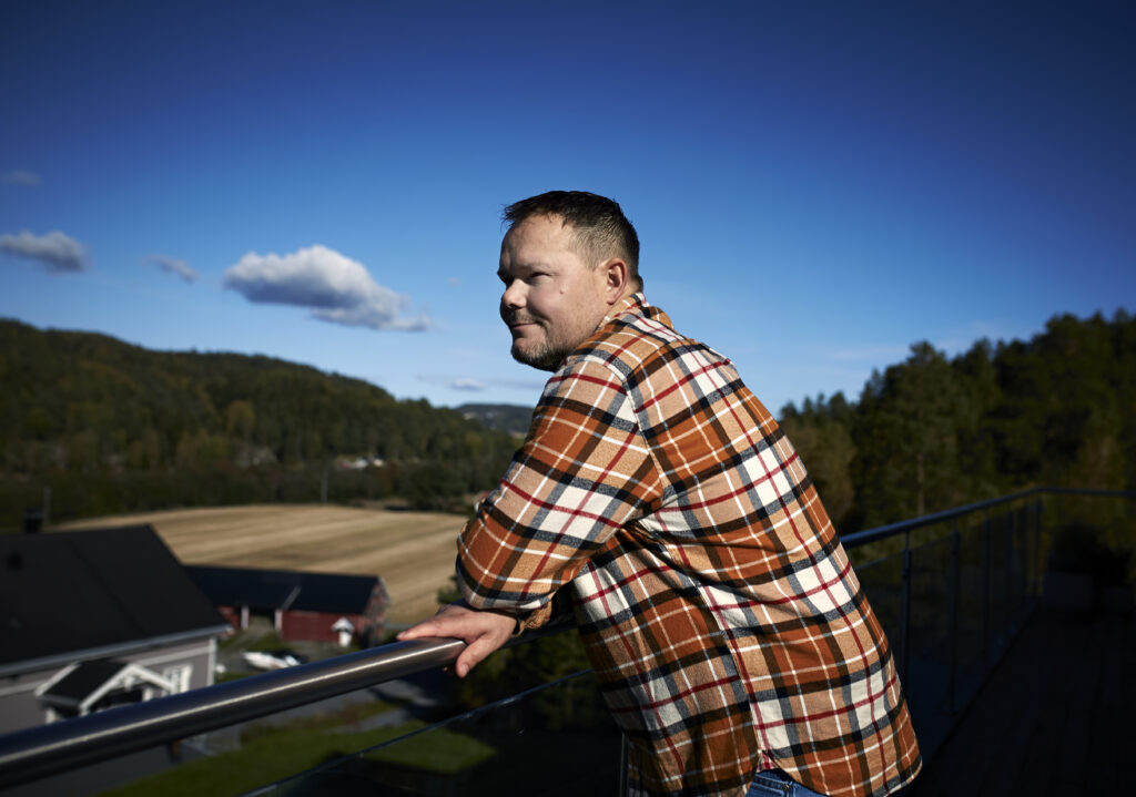 Mann i rutete skjorte lener seg på et rekkverk, med utsikt over et naturskjønt landskap med åkre og trær under en klarblå himmel.