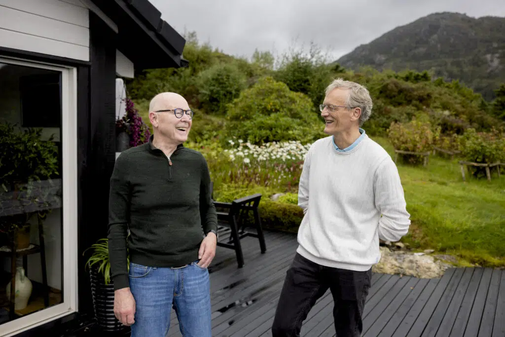 Ståle Myklebust og Halfdan Sørbye som smiler og snakker på et treterrasse utenfor et hus, med grønne åser og overskyet himmel i bakgrunnen.