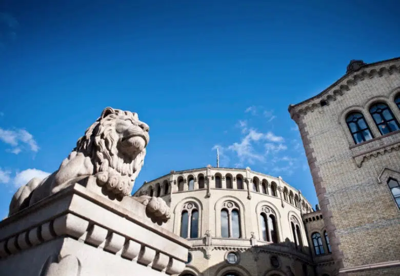 En løvestatue i stein foran en historisk murbygning med buede vinduer og blå himmelbakgrunn.