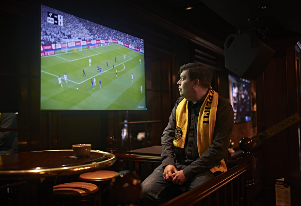 En mann med gult fotballskjerf ser på en fotballkamp på en stor skjerm i en svakt opplyst bar.