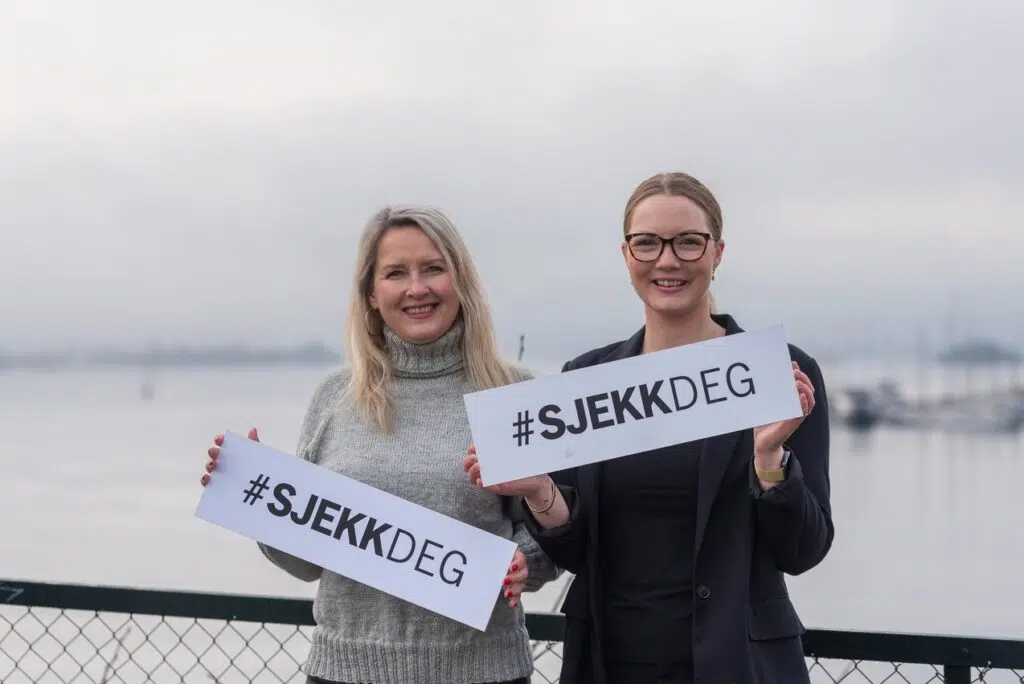To personer som holder «#SJEKKDEG»-skiltene utendørs nær en vannmasse. Bakgrunnen er tåkete, med båter synlige i det fjerne.
