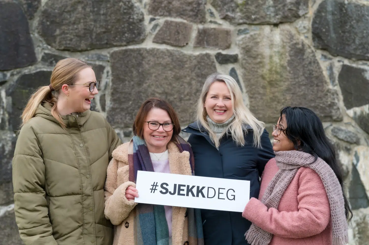 Fire personer smiler og holder et skilt med hashtaggen «#SJEKKDEG» foran en steinmur.