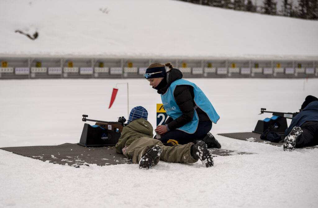 En person hjelper et barn med å sikte en rifle i snødekte utendørs omgivelser, med nummererte mål i bakgrunnen. Begge har på seg vinterklær.