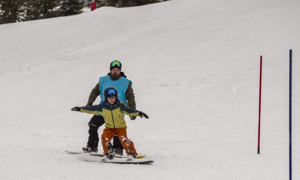 Voksen og barn på ski sammen i en snødekt bakke, begge iført blå vester og hjelm.