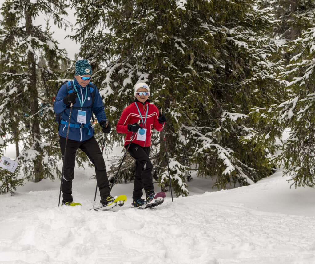 To personer på truger i en snødekt skog, iført vinterutstyr og solbriller.