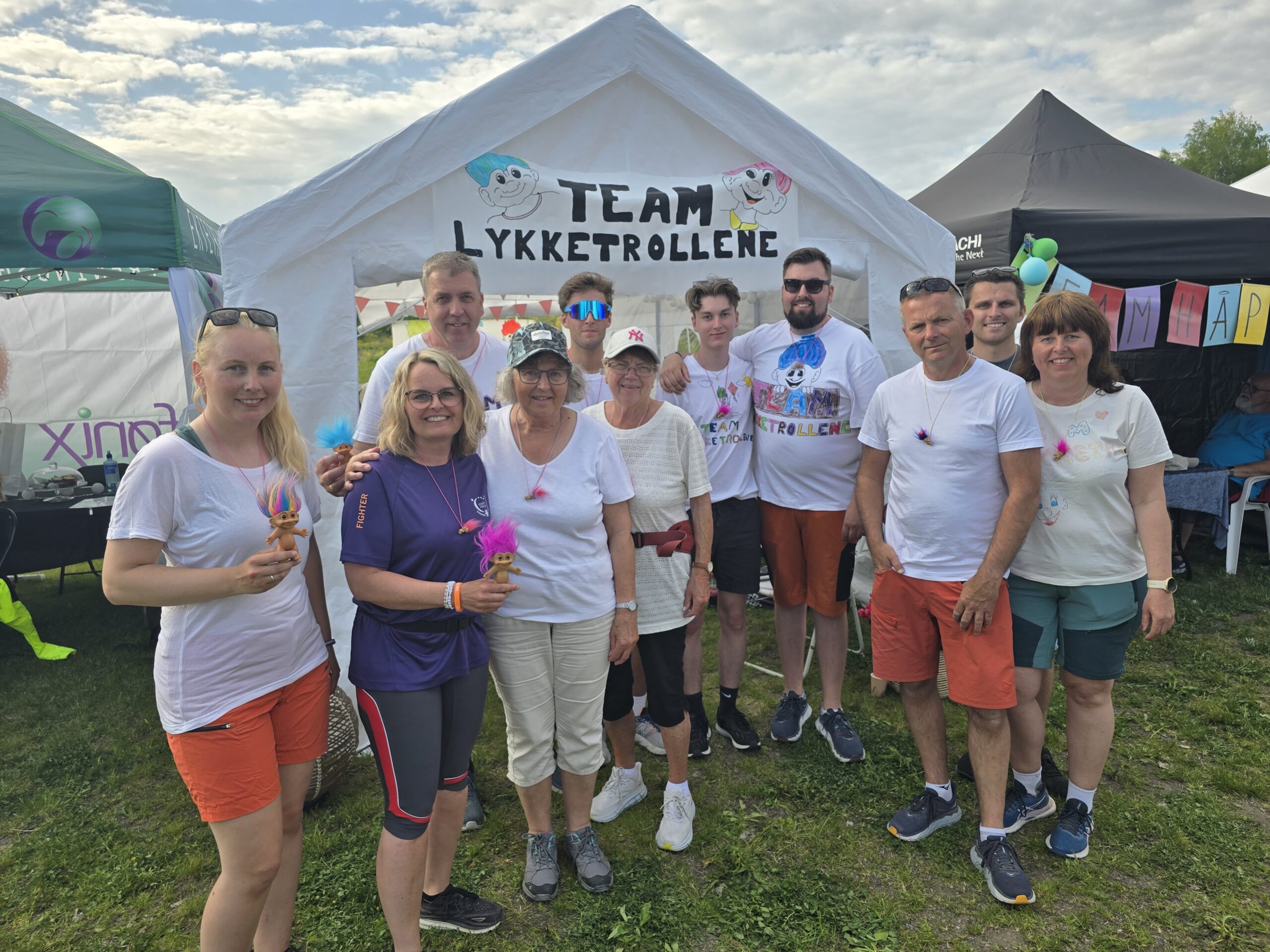 En gruppe mennesker poserer foran et telt med skiltet «Team Lykketrollene». De har på seg fritidsklær, stående på gresset, med andre telt og en delvis overskyet himmel i bakgrunnen.