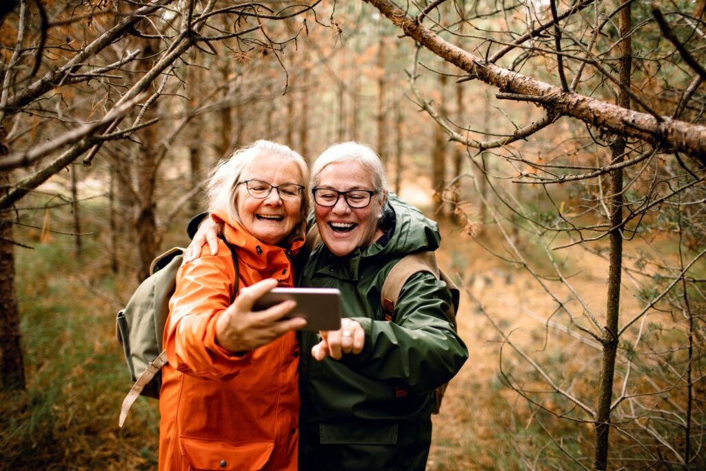 To eldre voksne iført jakker og ryggsekker tar en selfie mens de smiler i et skogsområde.