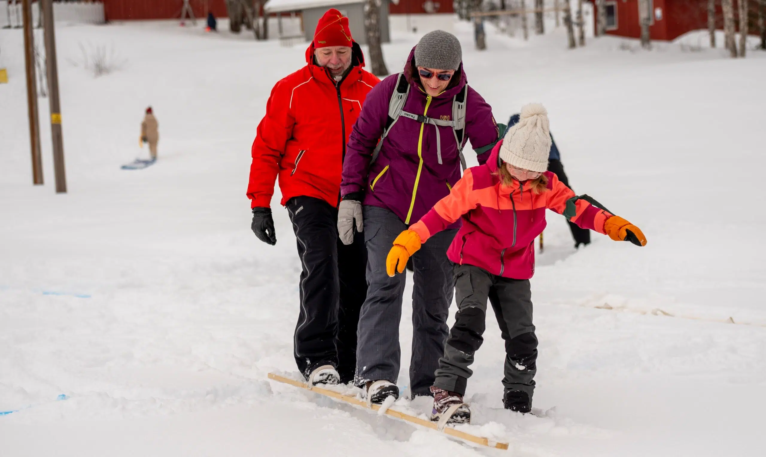 Tre personer som balanserer på en treplate i snøen, kledd i vinterklær, med et snølandskap og røde bygninger i bakgrunnen.