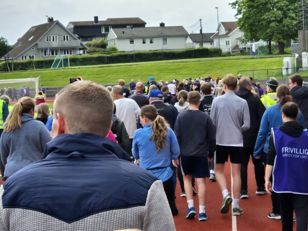 En stor gruppe mennesker som går på en bane i nærheten av bolighus, med noen iført fritidsklær og sportsklær.