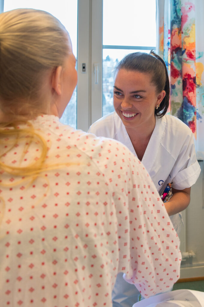 En helsepersonell i hvit uniform smiler mens han samhandler med en sittende pasient på sykehus.