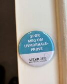 En knapp med norsk tekst "SPØR MEG OM LIVMORHALS-PRØVE," som betyr "Spør meg om cervical screening," og logo "SJEKK DEG KREFTFORENINGEN," på en hvit overflate.