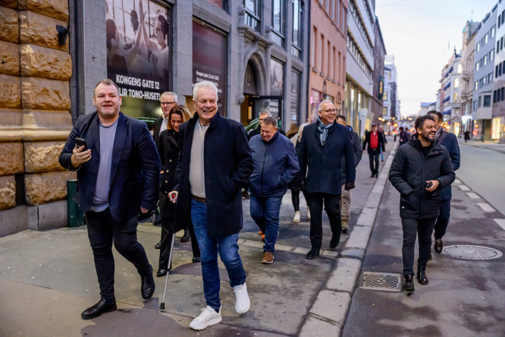 En gruppe mennesker, inkludert en person med krykker, går langs en bygate med bygninger.