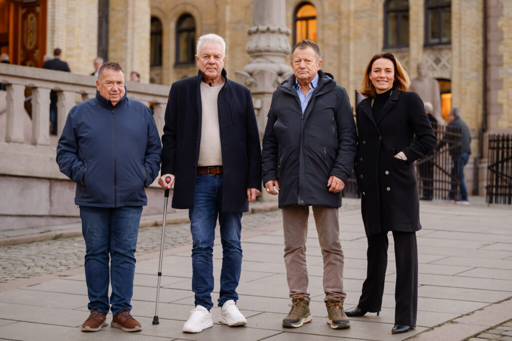 Fire personer, kledd i vinterantrekk, står på et steinfortau i urbane omgivelser, med en bygning i bakgrunnen. En person holder en spaserstokk.