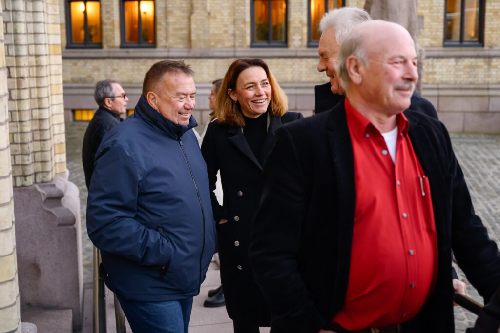 En gruppe mennesker står utenfor en bygning, kledd i vinterfrakker, engasjert i samtale.