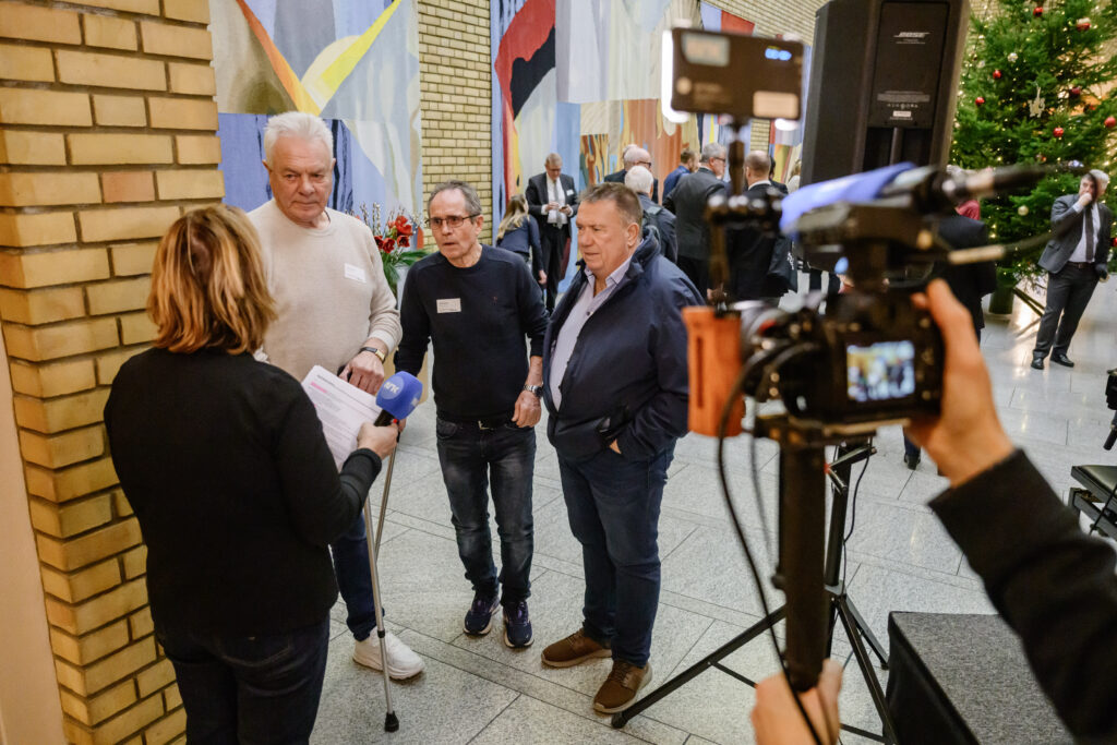 En journalist intervjuer tre menn i en gang. En kameraperson filmer intervjuet. Et juletre og store malerier er synlige i bakgrunnen.