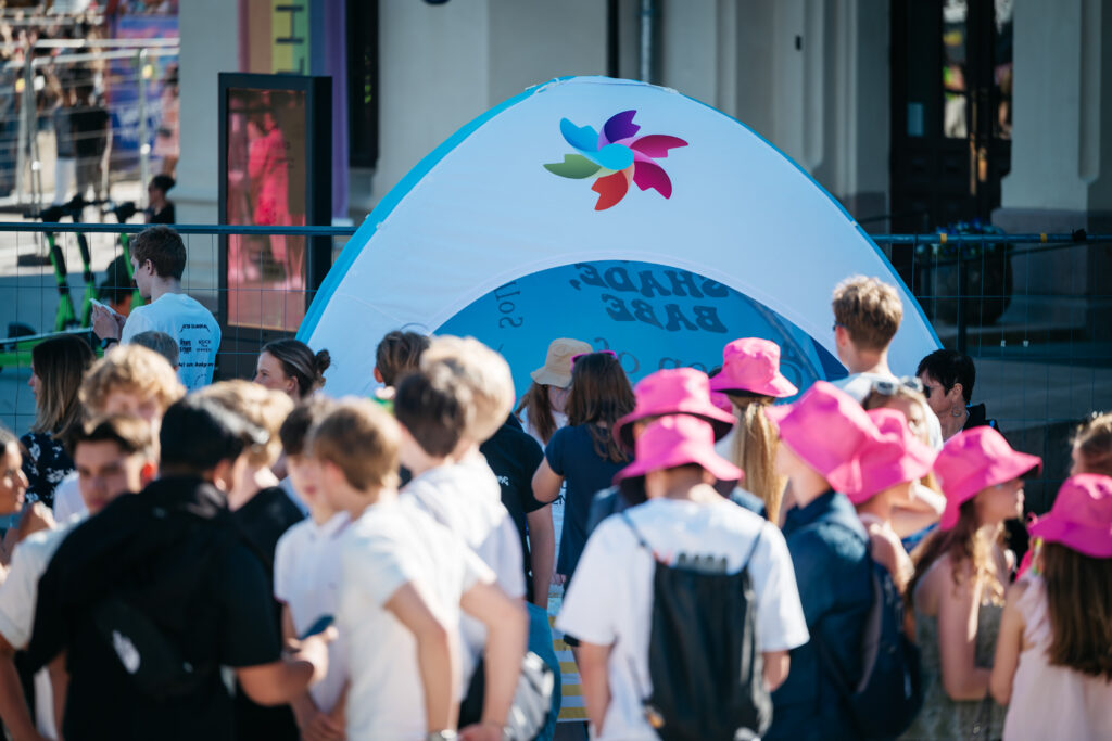 En mengde mennesker, noen iført rosa hatter, samles foran et telt med en fargerik logo.