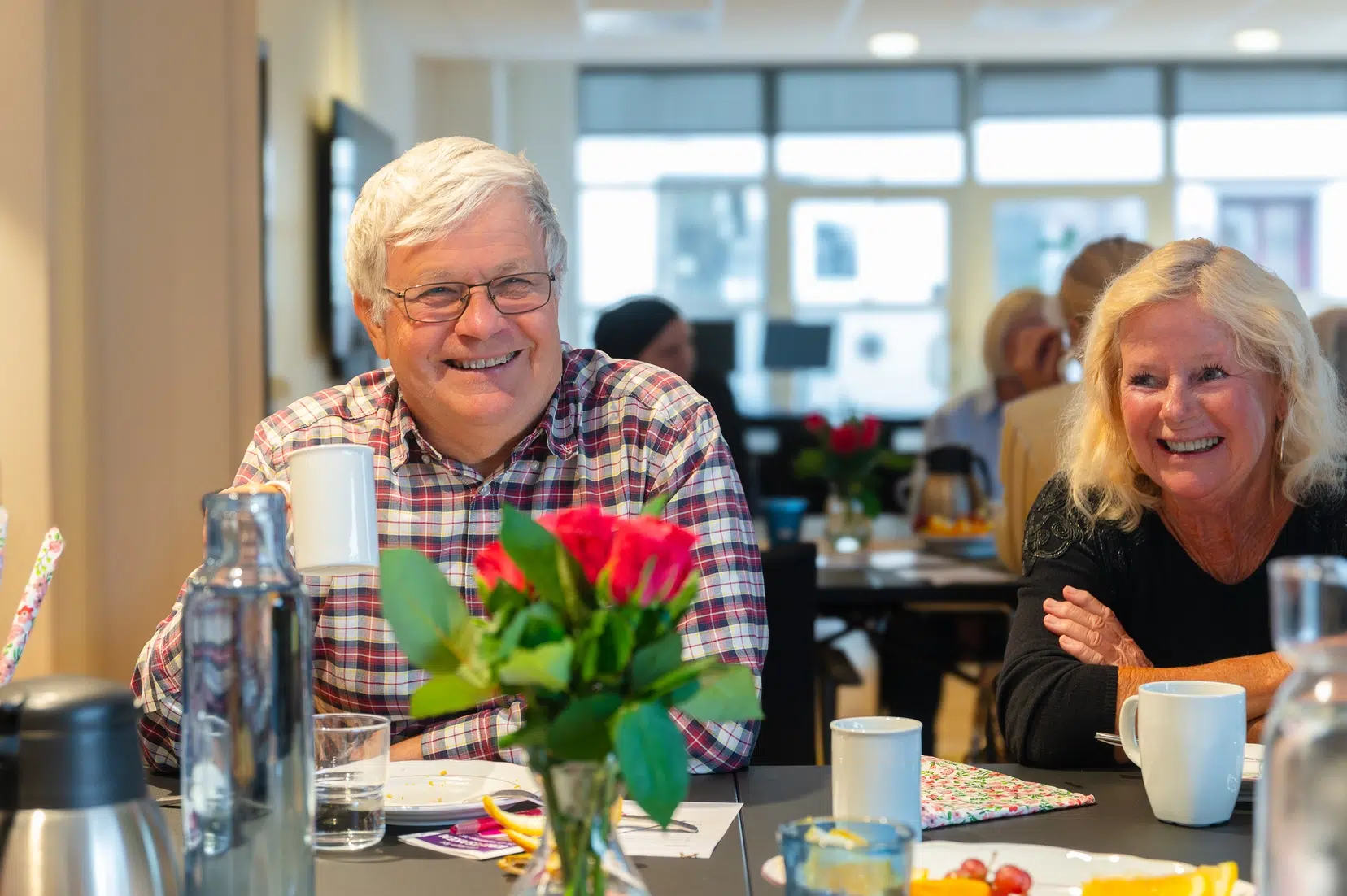 To personer som smiler og drikker kaffe ved et bord, med en vase med roser og forskjellige gjenstander rundt seg, i et sterkt opplyst rom.