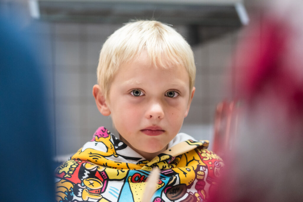 Små barn med kort blondt hår iført en fargerik hettegenser, ser direkte på kameraet.