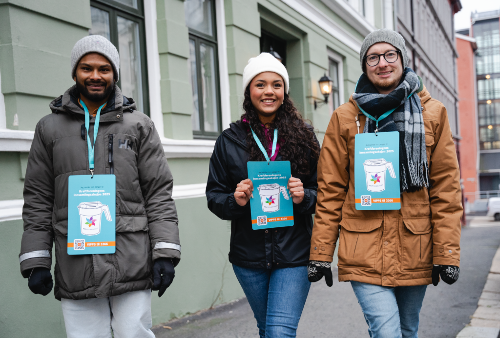 Tre personer i vinterklær står utendørs og smiler, holder skilt med en donasjonsrelatert melding, i urbane omgivelser.