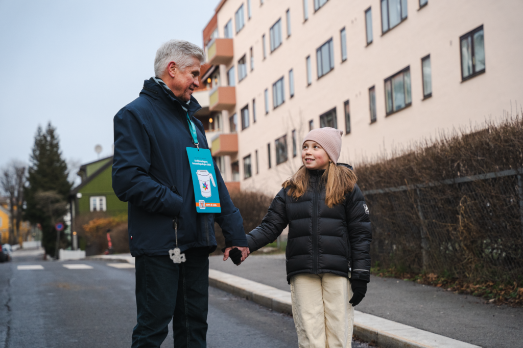 En eldre herre og et barn holder hverandre i hpnden, de står på en åpen gate med  blokker bak seg. Den eldre herren har på seg en innsamlingsplakat. 