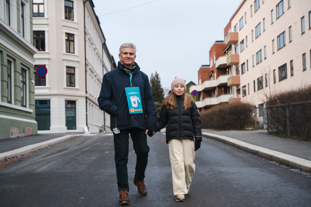 EN eldre mann og en jente holder hender og går nedover en gate med blokker bak. Den eldre herren har på seg en innsamlingsplakat.