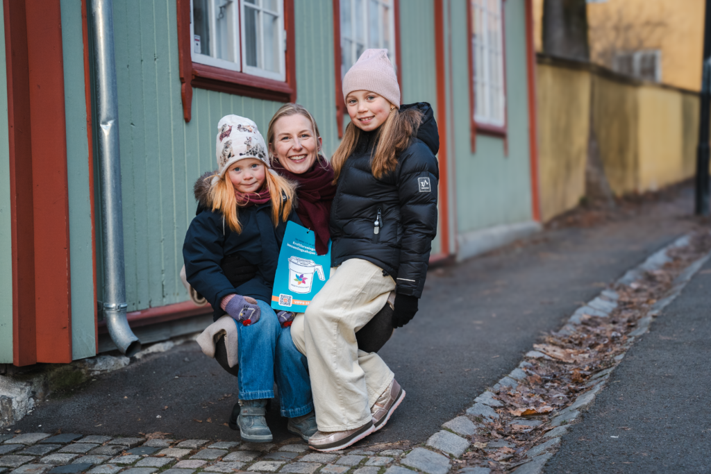 To barn sitter på fanget til en dame. De sitter utenfor et grønt hus og har på seg ytterklær, alle smiler og ser i kamera.