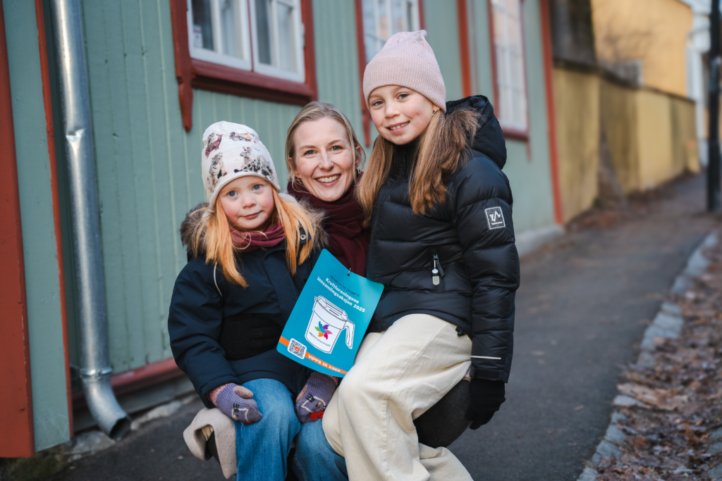 To barn sitter på fanget til en dame. De sitter utenfor et grønt hus og har på seg ytterklær, alle smiler og ser i kamera. 