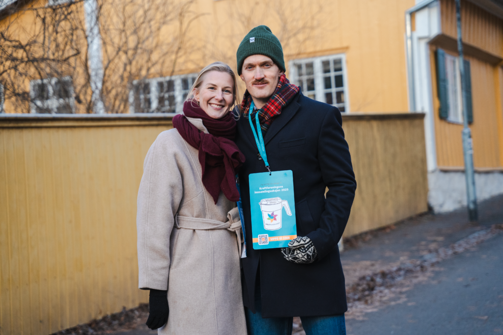 En kvinne og en mann holder rundt hverandre og ser inn i kamera og smiler, De står ute foran et gult gjerne og smiler til kamera. begge har på seg innsamlingsmateriell. 