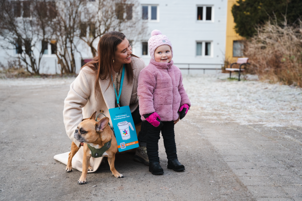 En kvinne sitter på huk ved sin av en jente i rosa teddyjakke. Ved siden av kvinnen sitter det en liten hund. 