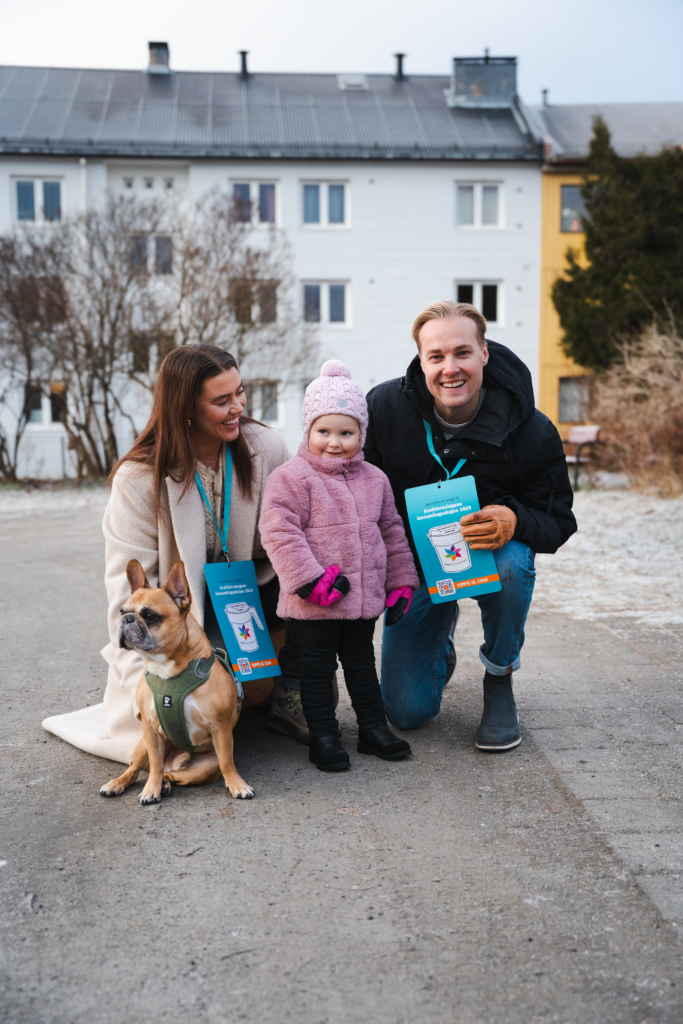 En kvinne, et barn og en mann står på en gårdsplass og smiler til kamera. Ved siden av kvinnen er det en hund.