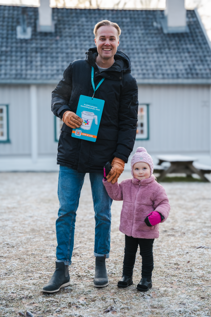 En mann og et barn står på en gårdsplass og har på sge vinterklær. Begge er i kamera og smiler, mannen holder en innsamlingsplakat. 