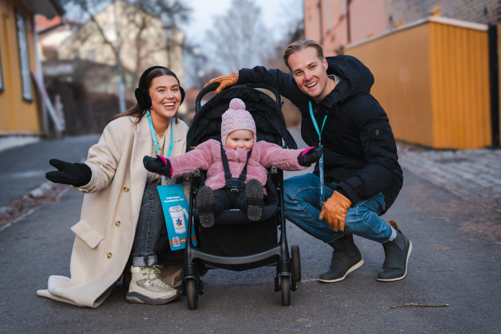 En kvinne og en mann sitter på huk på hver sin side av en barnevogn med en jente som smiler til kamera.