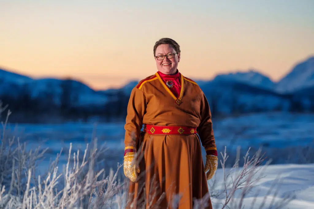 En person i tradisjonell drakt står utendørs i et snødekt landskap med fjell i bakgrunnen under solnedgang, som symboliserer hvordan HPV hjemmetest gir råderett over egen kropp.