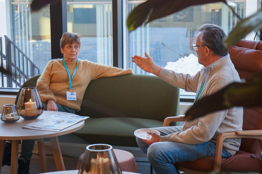 To personer sitter i et moderne salongområde, engasjert i samtale. En person gestikulerer mens han snakker. Et bord med et stearinlys og papirer er i forgrunnen.