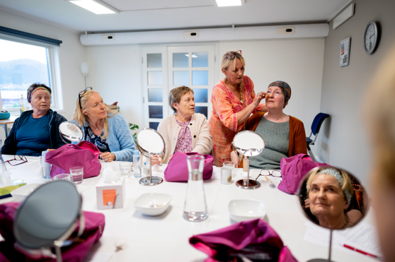 En gruppe kvinner deltar på et sminkeverksted, sittende rundt et bord med speil og kosmetikk, mens en instruktør demonstrerer teknikker på én deltaker.