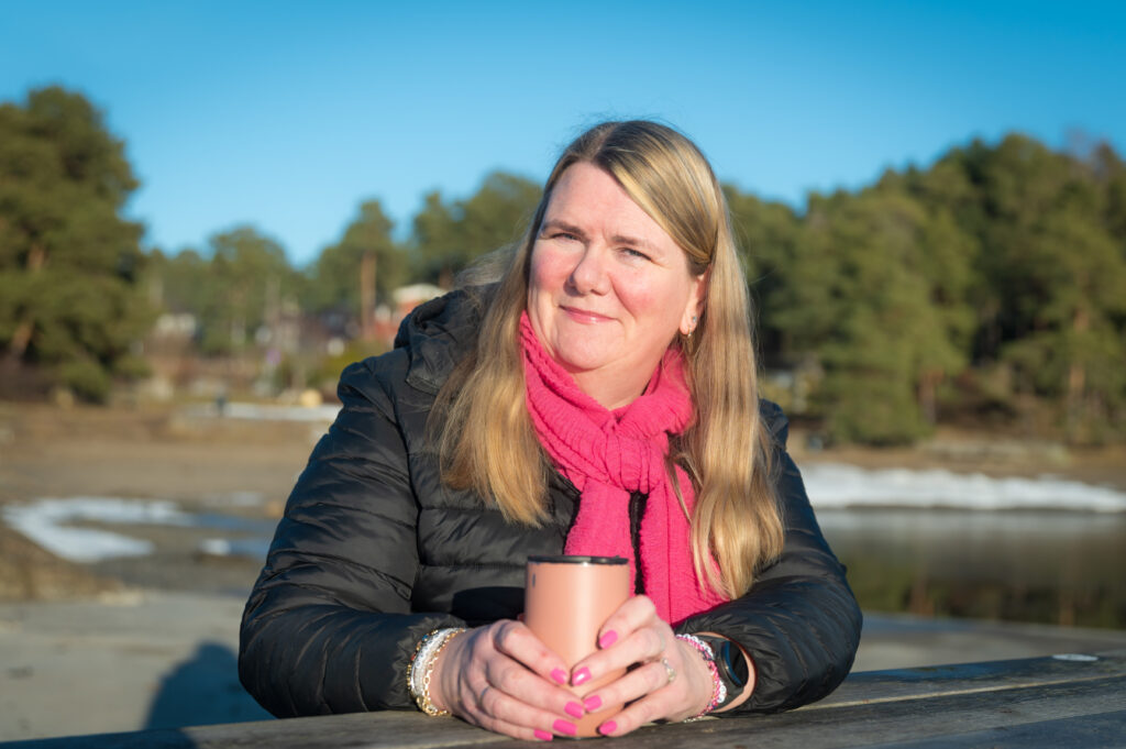 En kvinne med langt hår, iført svart jakke og rosa skjerf, holder en kopp mens hun sitter utendørs ved et trebord, med trær og en elv i bakgrunnen.