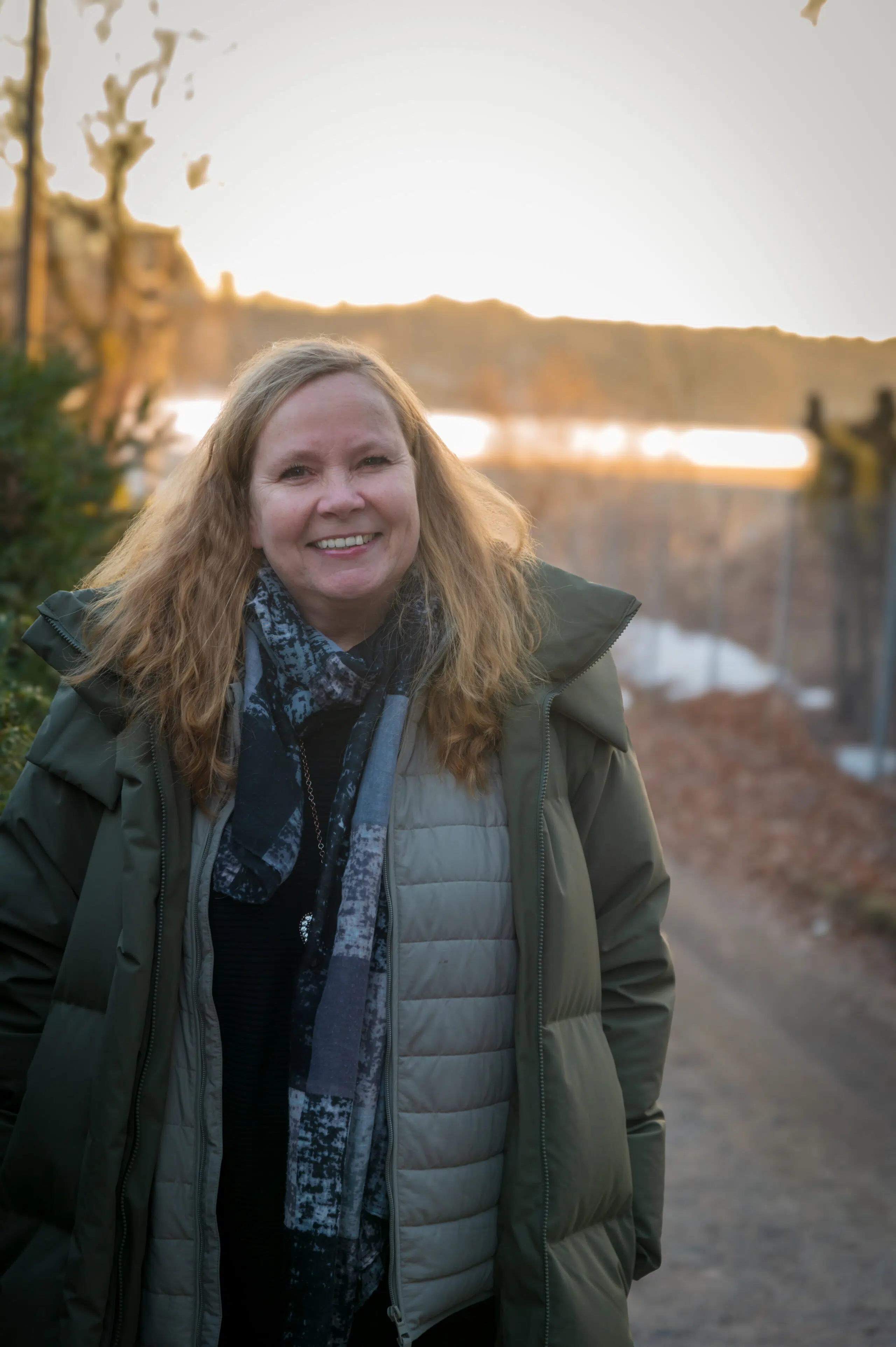 Person med langt hår, smilende, iført mørk frakk og skjerf, står på en sti med et solnedgangsopplyst landskap i bakgrunnen.