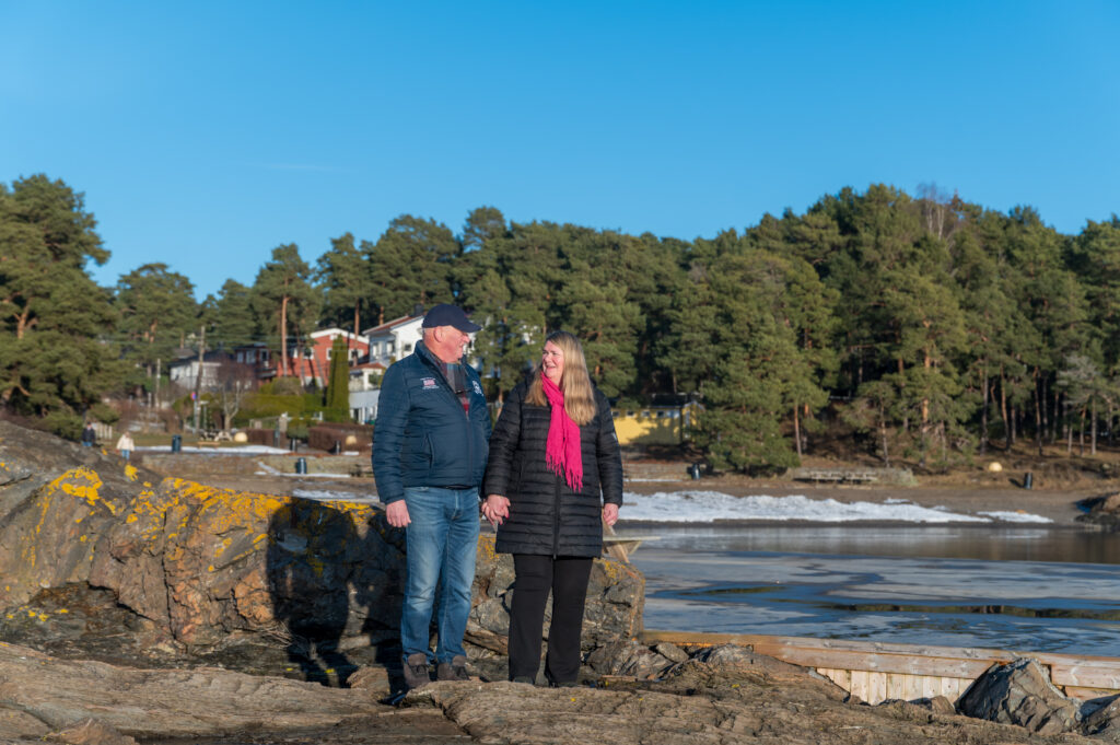 En mann og kvinne står på steinete terreng nær en frossen vannmasse, omgitt av trær og hus under en klarblå himmel.