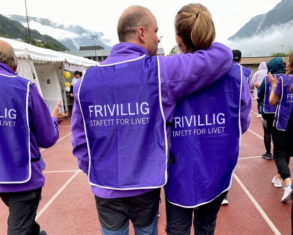 To personer iført lilla vester med «Frivillig Stafett for Livet» står arm i arm på en bane. Andre deltakere er i bakgrunnen.