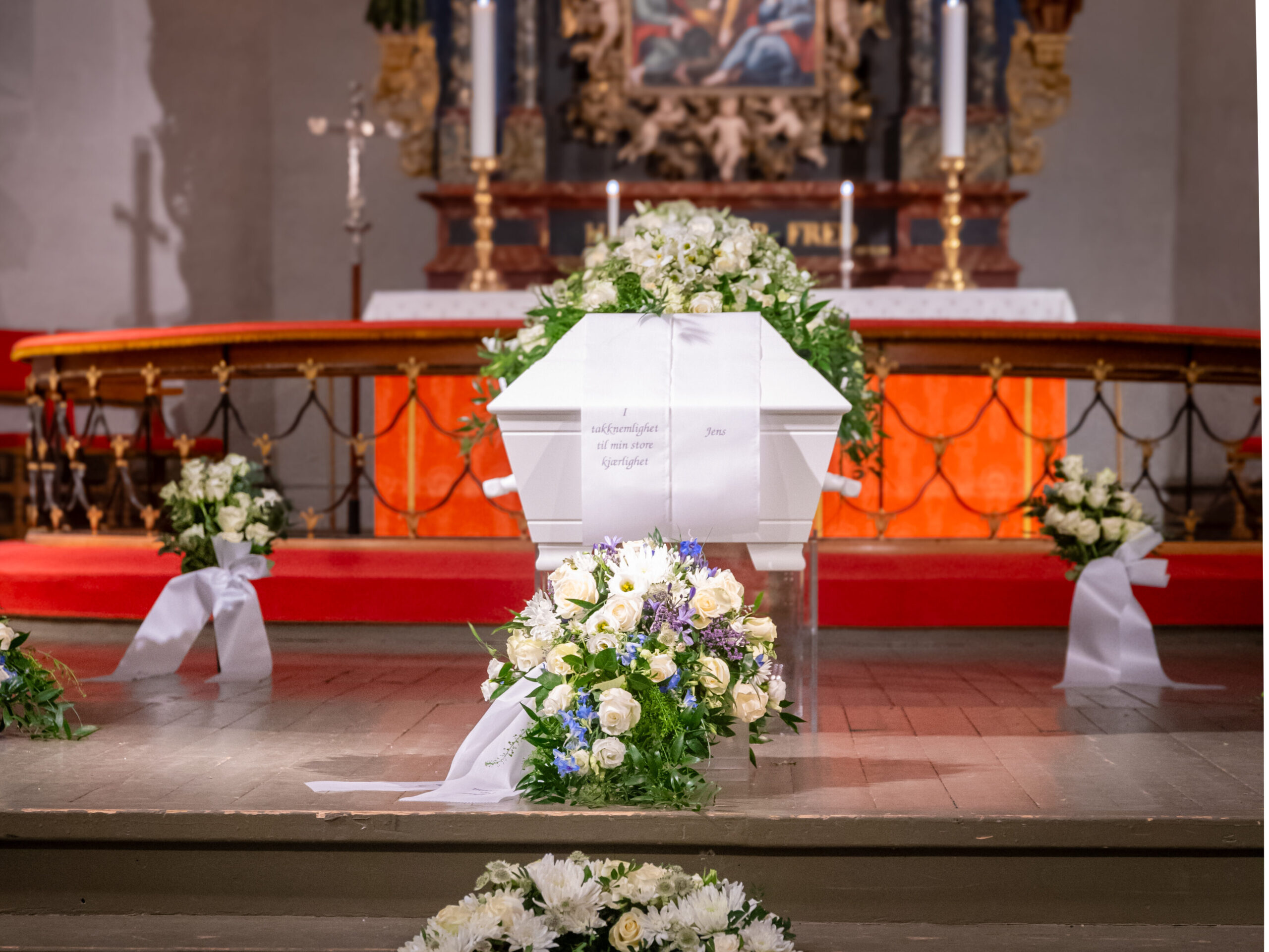 En hvit kiste pyntet med blomster i en kirke, omgitt av blomsteroppsatser og kranser på et tregulv.