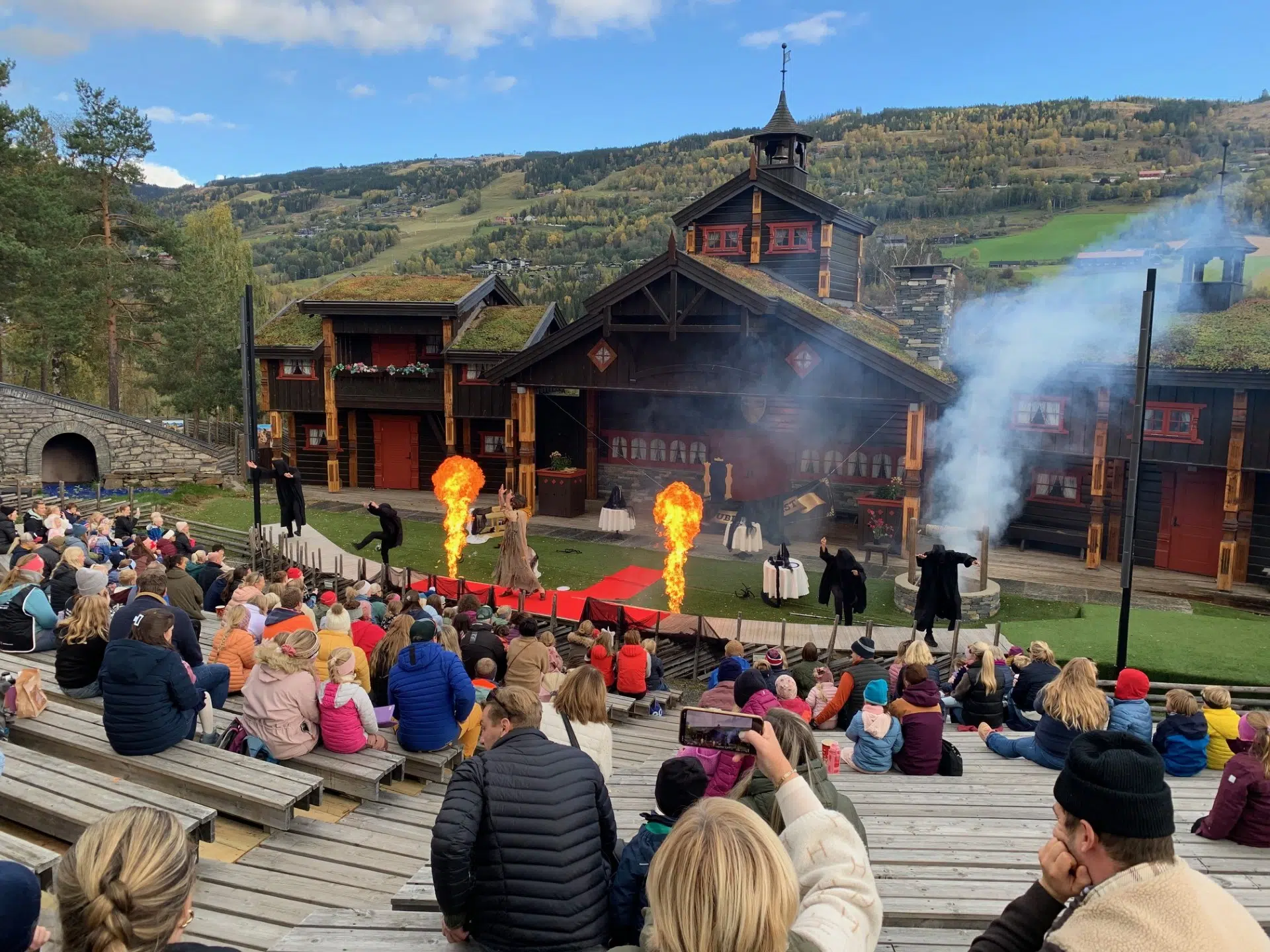 Utendørsforestilling med skuespillere som skaper branneffekter på en scene. Publikum som sitter på benker ser på showet. Naturskjønn bakgrunn med åser og trær.