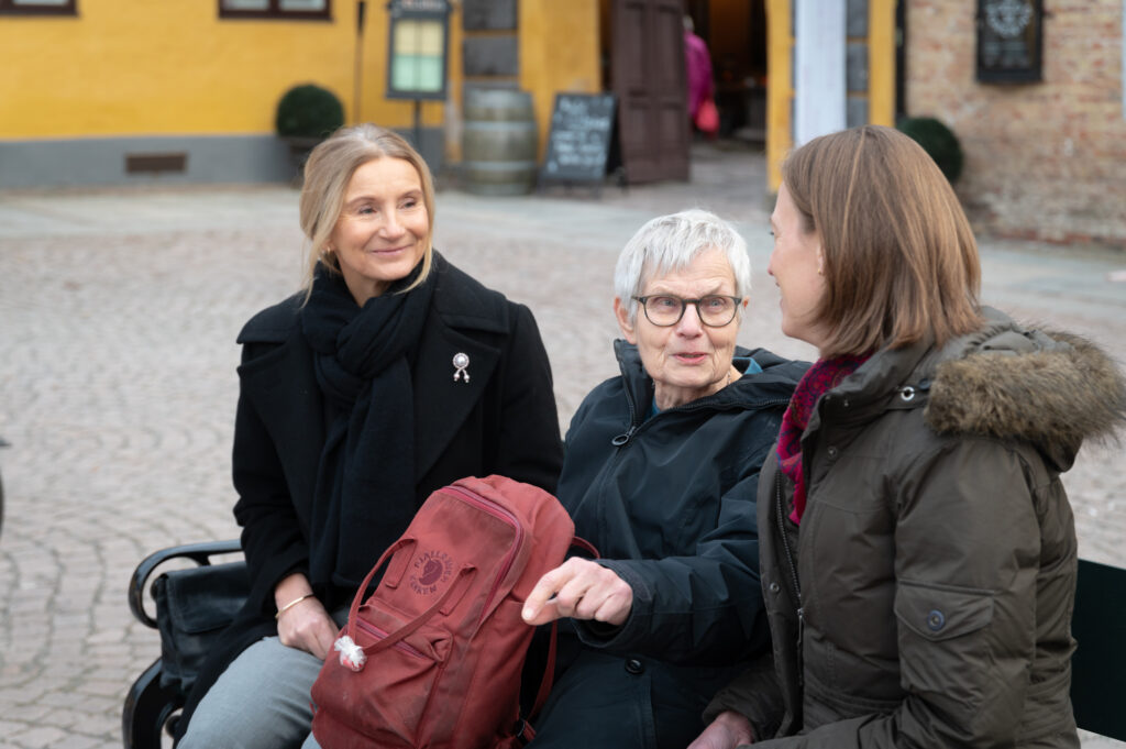 Tre kvinner sitter på en benk i en utendørs setting og deltar i samtale. En kvinne holder en rød ryggsekk. Bakgrunnen inkluderer en gul bygning og brosteinsgrunn.