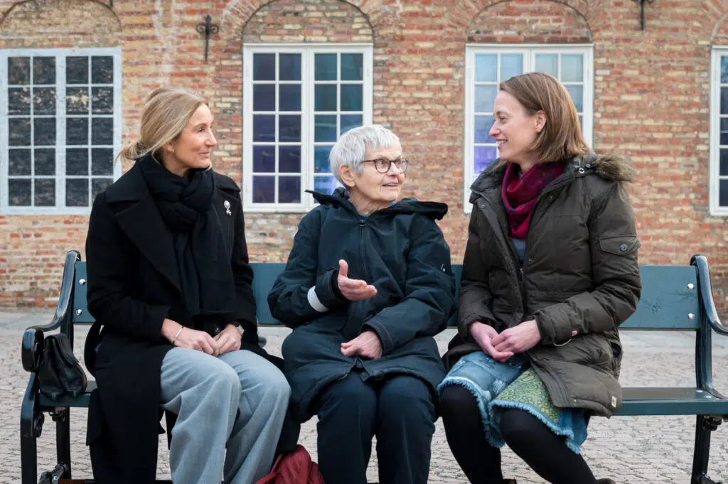 Tre personer sitter på en benk, engasjert i samtale. Det er en murbygning med vinduer i bakgrunnen.