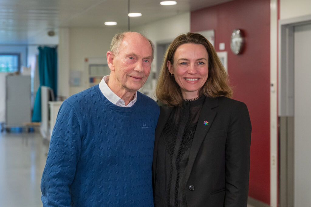 En eldre mann i blå genser står ved siden av en smilende kvinne i svart antrekk i en gang med røde og hvite vegger.