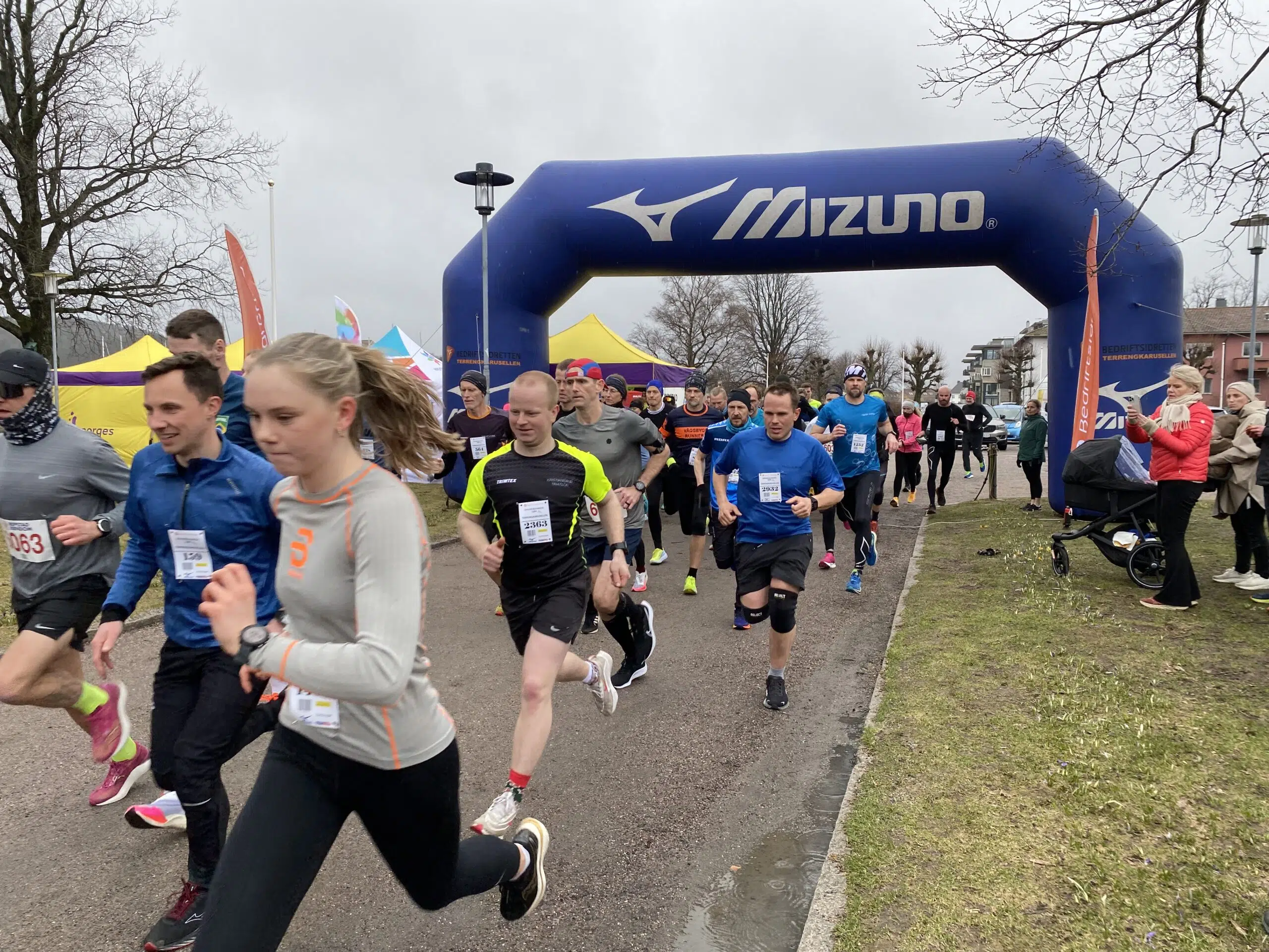 En gruppe løpere som deltar i et maraton, passerer under en blå oppblåsbar bue med "Mizuno" skrevet på. Tilskuere langs sidene, og været er overskyet.