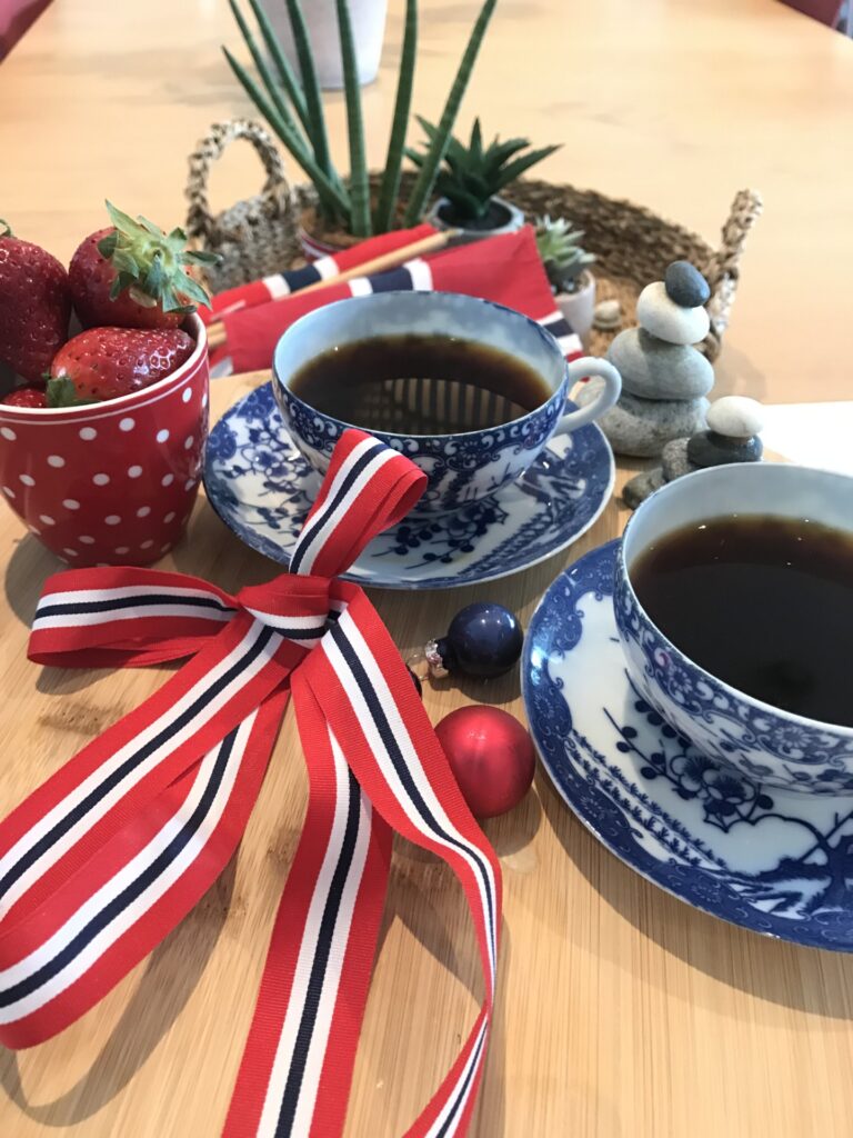 To blå og hvite tekopper med svart kaffe, en rød prikkete kopp jordbær, et rødt-hvitt-blått bånd og små steiner arrangert på et trebord.