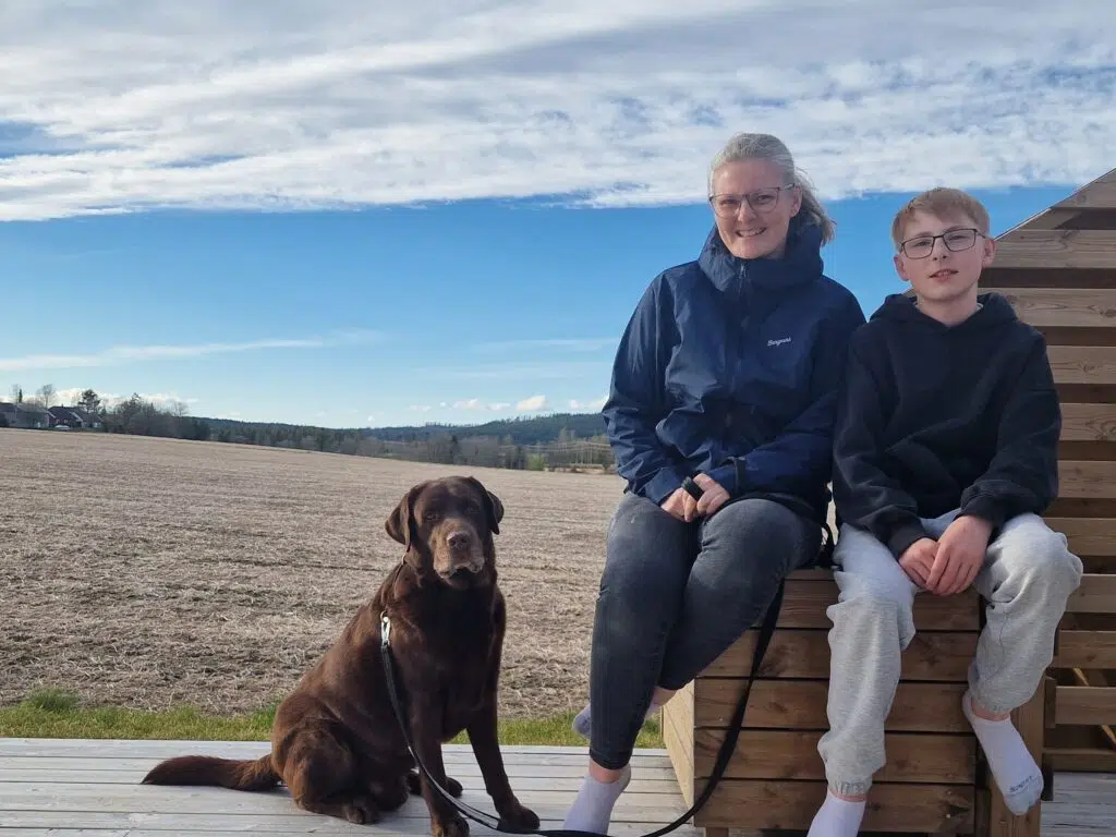To personer sitter på en treterrasse ved siden av en brun hund med åkre og en delvis overskyet himmel i bakgrunnen.