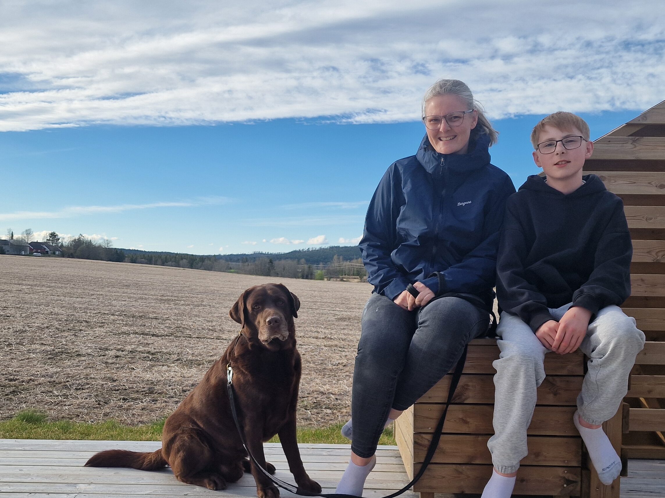 To personer sitter på en treterrasse ved siden av en brun hund med åkre og en delvis overskyet himmel i bakgrunnen.