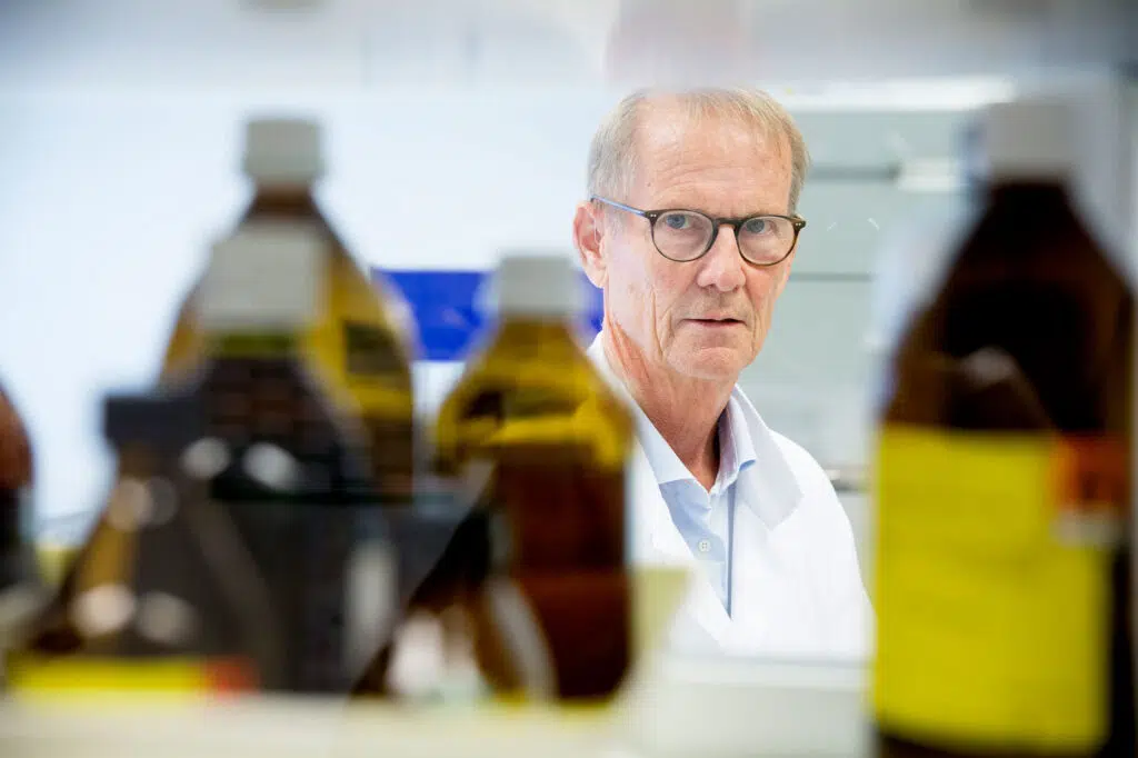 En person i en hvit laboratoriefrakk er omgitt av store brune flasker i laboratoriemiljø.