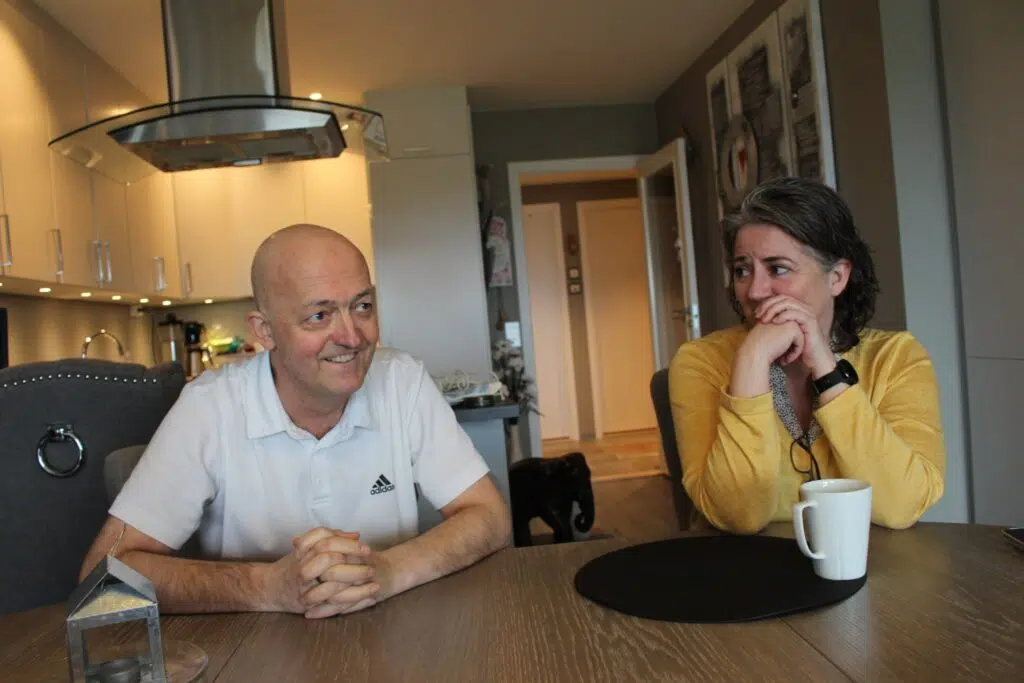 En mann og en kvinne sitter ved et kjøkkenbord, vendt mot hverandre. Mannen smiler med hendene foldet, mens kvinnen, kledd i gult, ser på ham med haken hvilende på hånden.